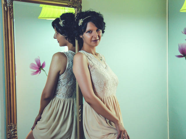 AmethystCharm stands in front of a mirror, wearing a lace sleeve dress, with curly black hair and a flower headpiece, her reflection showing her soft tits.