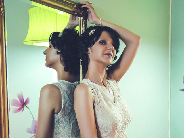 AmethystCharm with curly black hair, wearing a white lace sleeveless top, arms raised, reflecting in a mirror, green lamp in background, pink flower, and a purple necklace.