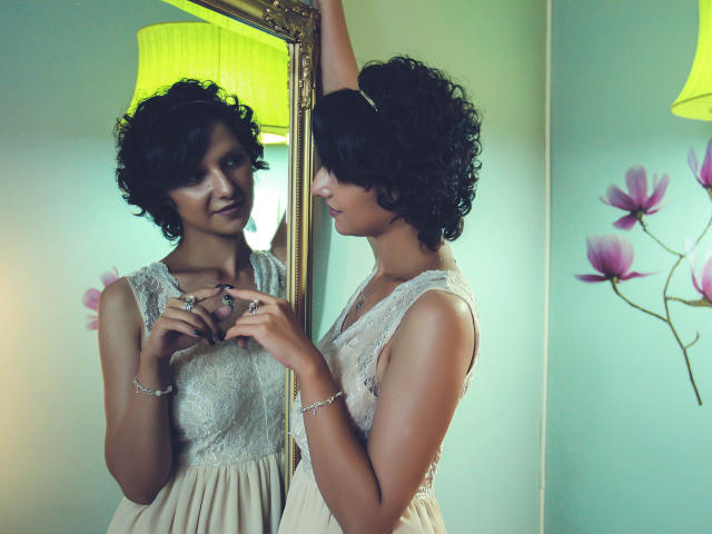 AmethystCharm with curly black hair, wearing a sleeveless lace dress, standing in front of a mirror, adjusting a ring, reflecting her face and small tits in the mirror.
