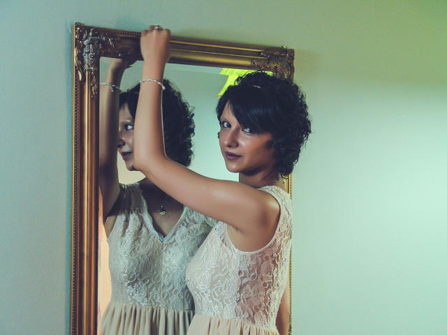 AmethystCharm with curly black hair, wearing a lace dress, stands in front of a mirror, holding the frame with her right arm, reflecting her serious, dark-eyed face.