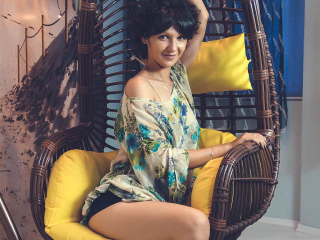 AmethystCharm with short black hair, smiling, wearing a floral shirt and black panties, sitting in a wicker chair with yellow cushions, one arm raised, showing her tits.