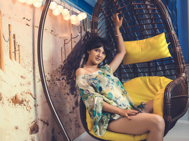 AmethystCharm with curly black hair, wearing a floral off-shoulder dress, sits in a wicker hanging chair with yellow pillows, her arm raised, exposing her bare, smooth-ass.