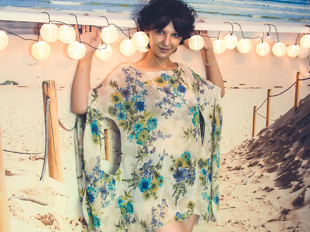 AmethystCharm with short curly black hair, wearing a floral sheer dress, standing in front of warm string lights, holding her dress, showing her tits through the fabric.