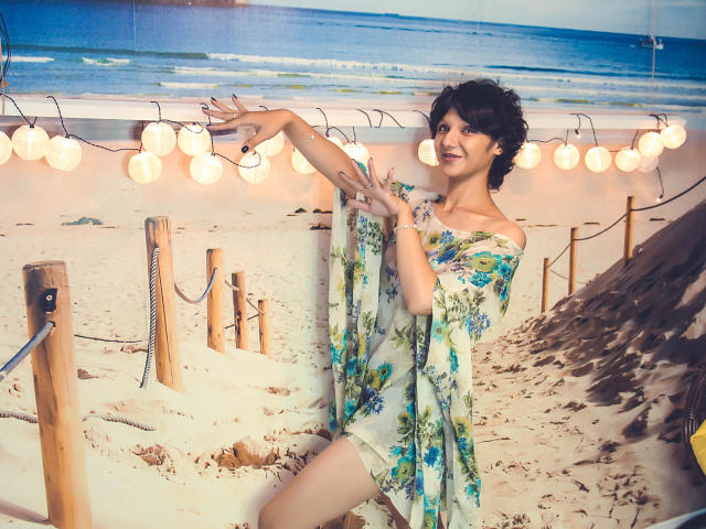 AmethystCharm with short curly black hair, wearing a floral off-shoulder dress, smiling and gesturing with hands raised. She's on a sandy beach with string lights.