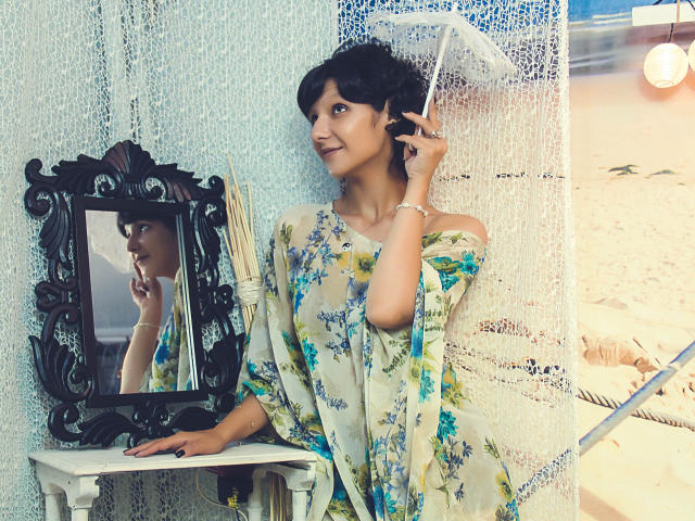 AmethystCharm with short black hair, floral dress, holding phone to ear, standing in front of ornate mirror on white table, lace curtain, light bulb, sandy beach in background.