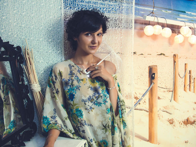 AmethystCharm with dark hair, wearing a floral shawl, holding a paintbrush, standing by a mirror, looking at the camera, smiling, showing her tits, against a textured wall.