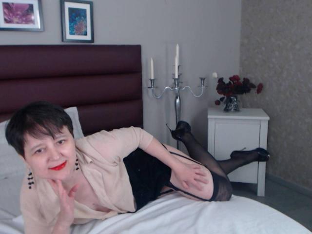 ChristaRose with short black hair, red lipstick, beige blouse, black garter, stockings, and heels, lying on bed, smiling, showing her tits and ass, near white nightstand with candles.