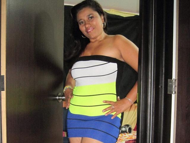 KatthyBabe con un sedere importante, sorridente, indossa un abito a righe senza spalline con colori nero, bianco, giallo e blu, in piedi in una porta, capelli scuri, orecchini a cerchio, mani sui fianchi..