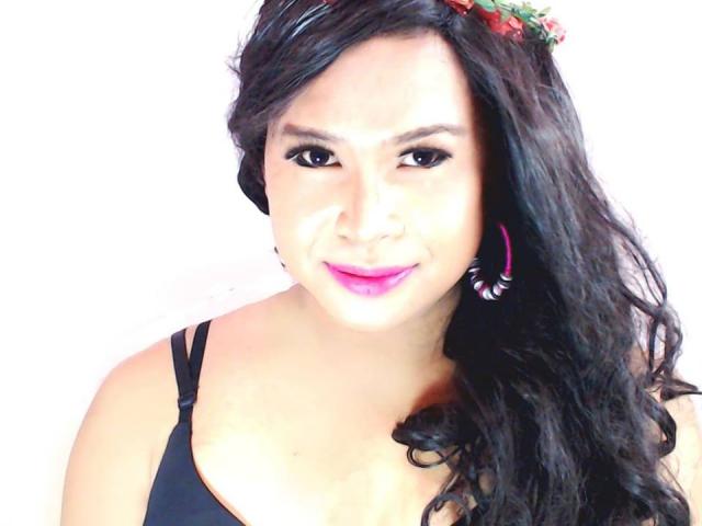 Sheena Collins hat schwarzes, welliges Haar, pinken Lippenstift und trägt ein schwarzes Oberteil mit pinken Ohrringen. Ihr Gesicht ist leicht lächelnd, und sie hat eine kleine rote Blume in ihrem Haar..