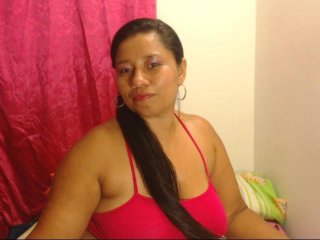 KatthyBabe tiene la piel morena, cabello negro largo, un top rojo que deja ver sus senos, maquillaje de ojos rosa, aros grandes y está sentada con una cortina rosa detrás de ella..
