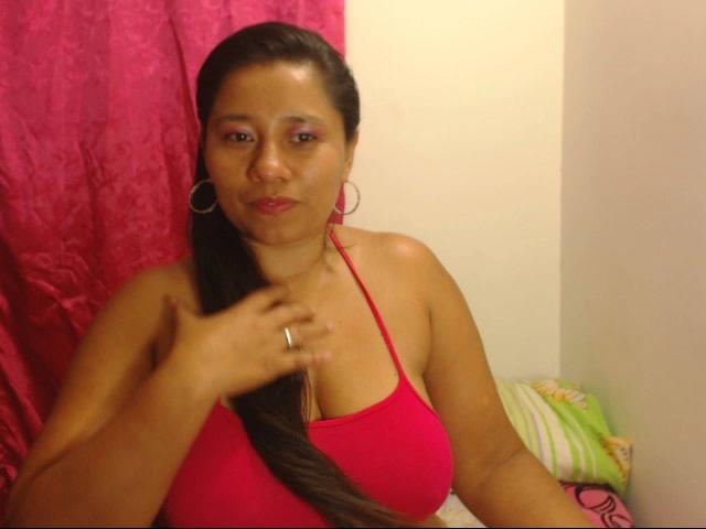 KatthyBabe con lunghi capelli castani, top rosso che lascia intravedere il seno, orecchini a cerchio rosa, anello alla mano destra, espressione seria, tenda rosa sullo sfondo, seduta sul letto..