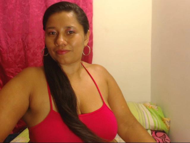 KatthyBabe with medium brown skin, red halter top, showing her large tits, long black ponytail, pink lipstick, hoop earrings, sitting on bed with green and pink pillows.