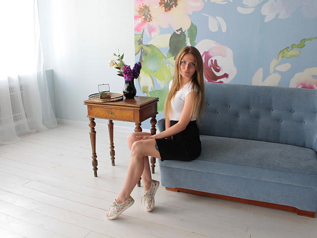 GretyArdent está sentada en un sofá azul, vestida con una blusa blanca, una falda negra y zapatos blancos, con las piernas cruzadas, mostrando sus glúteos, y un jarrón con flores moradas sobre una mesa..