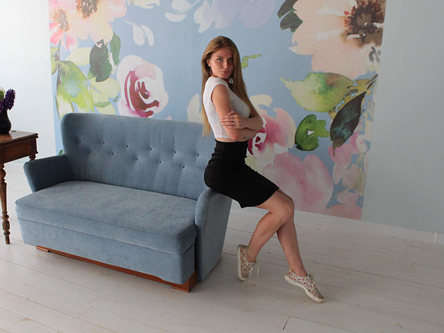 GretyArdent, avec de petites seins, assise sur un canapé bleu, portant un crop top blanc, une jupe noire et des baskets blanches, les bras croisés, dans une pièce avec du papier peint floral et un sol blanc..