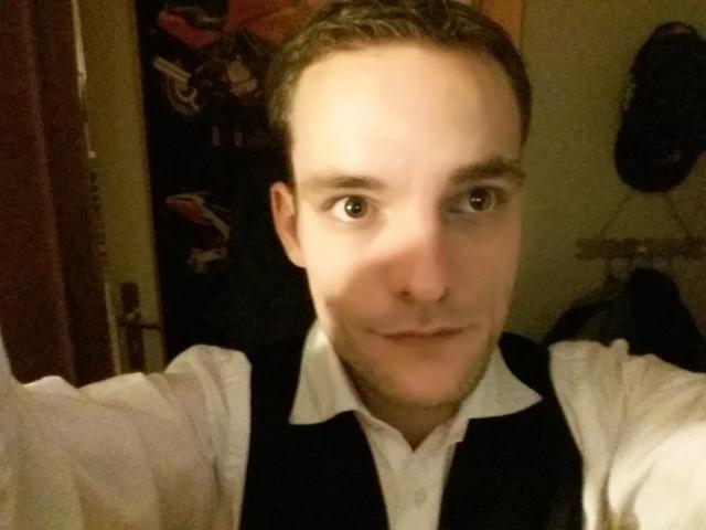 Dildoxxlhard, avec des cheveux bruns courts, une peau claire et des yeux bruns, portant une chemise blanche et un gilet noir, souriant à la caméra. Éclairage intérieur chaleureux..