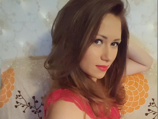 ToiSecret has long brown hair, red lipstick, and is wearing a red lace top, looking over her shoulder with a slight smile. Her tits are partially visible.