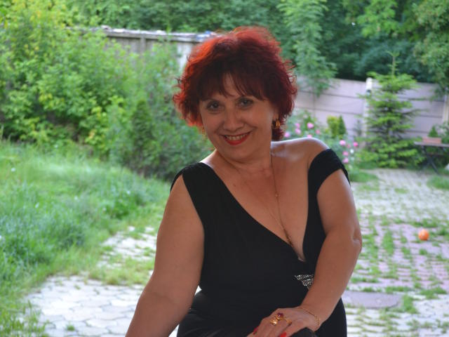 SexyEllaForYou with red curly hair, black dress with deep cleavage, smiling, showing her boobs, standing in a grassy garden, holding a bike, wearing a ring.