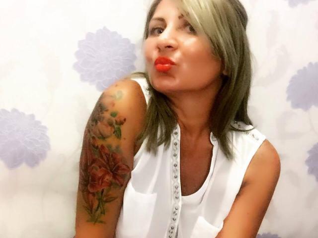 ChaudeEvely with red lips, pouty expression, and blonde-green hair, wearing a white sleeveless top, showcasing a colorful rose tattoo on her right arm.