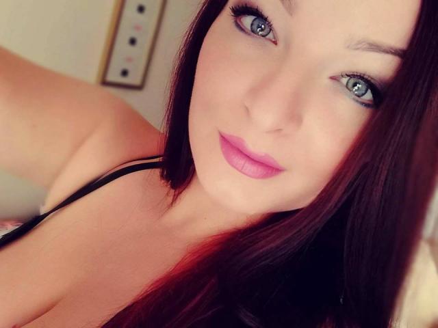 StellaLee heeft blauwe ogen, roze lippenstift en lang bruin haar, en draagt een zwarte top met bandjes, waarbij haar decolleté zichtbaar is. Haar make-up is subtiel, en ze glimlacht lichtjes..