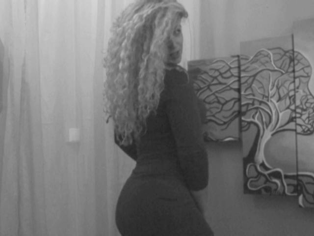 SassyKate with curly blonde hair, wearing a tight dark dress, showing her ass, standing in a room with abstract tree paintings on the wall. Black-and-white photo.