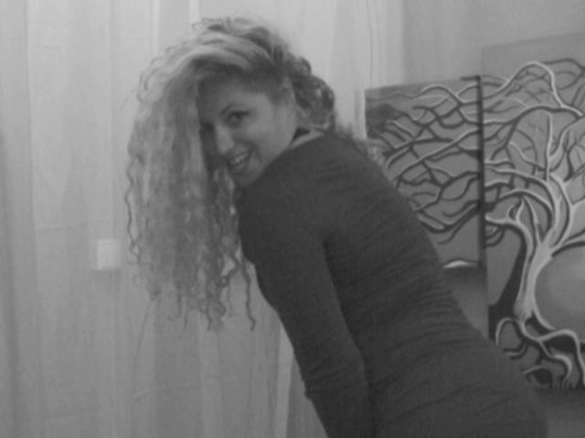 SassyKate with long curly hair, smiling at the camera, wearing a tight, dark, long-sleeve top, bending slightly, showing a cute, playful, and confident look. Black-and-white photo.