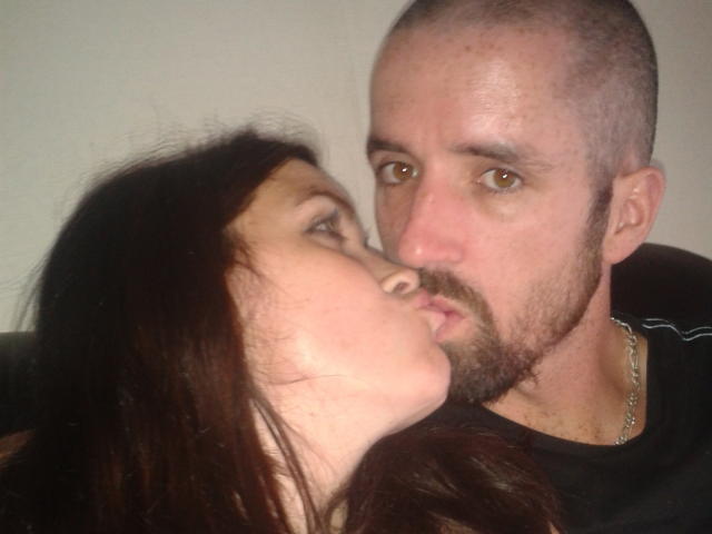 Un couple, dont l'un est chauve et a une barbe, avec des yeux marron, embrasse une femme brune sur la joue. Les deux se regardent, la bouche légèrement ouverte..
