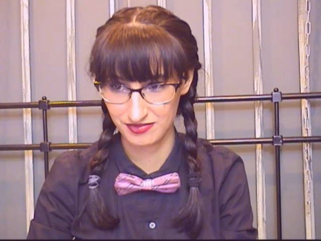 LysAddiction has brown hair in braids, glasses, red lipstick, black shirt, pink bowtie, and is looking down with a slight smile. Her tits are small.