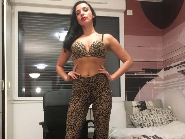 KatieFrenchie con un sujetador y pantalones de estampado de leopardo, de pie, con las manos en las caderas, lápiz labial rojo, cabello negro, senos medianos, dormitorio, silla negra, ventana con persianas, cama con almohadas..