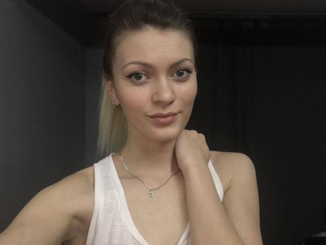BlossomPussy, ein süßes Mädchen mit kleinen Brüsten, heller Haut und einem blonden Pferdeschwanz, trägt ein weißes Tanktop, hält sich am Hals und lächelt sanft, und trägt eine silberne Halskette..