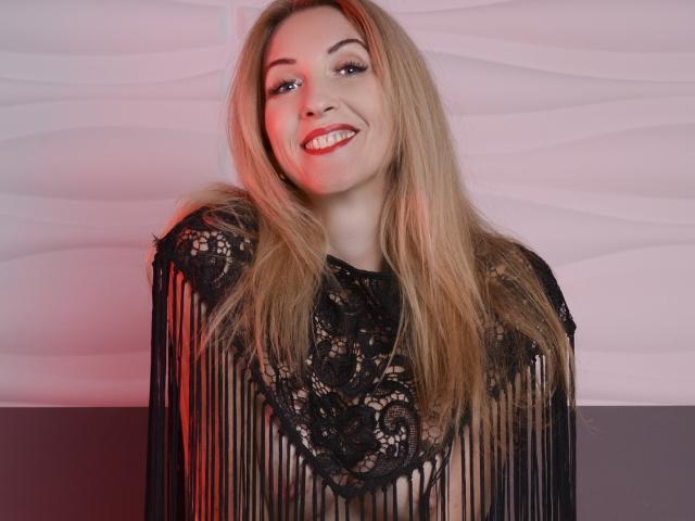 LadyLea, con lunghi capelli biondi, sorridente, indossa una maglia nera in pizzo con frange, che lascia intravedere i suoi seni nudi attraverso il pizzo. È attraente, con un viso carino e rossetto rosso..