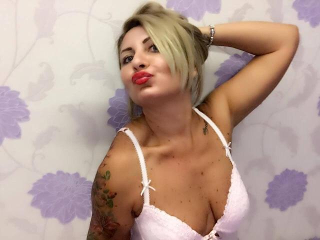 ChaudeEvely with blonde hair, puffy red lips, white lace bra, tattoos on arm, hand in hair, looking up, showing boobs, pink skin, bracelet on wrist.