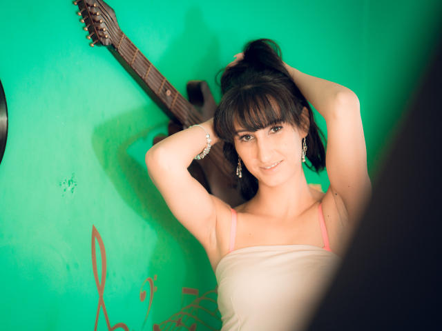 LysAddiction with dark hair, smiling, wearing a white strap top, arms raised, holding hair, green wall, acoustic guitar, earrings, and a bracelet. Boobs visible.
