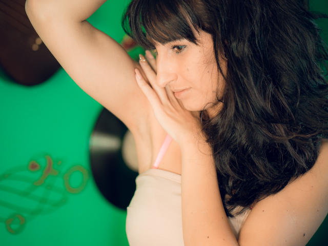 LysAddiction with dark wavy hair, pale skin, and small tits, lifting her arm, showing a pink bra, hand on her armpit, green background, slightly smiling, upper body shot.