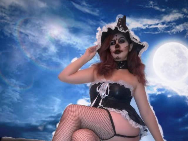 WonderPearl com maquiagem escura, cabelo ruivo, chapéu de bruxa preto, corpete de couro preto sem alças, meias de rede, sentada, mostrando o bumbum, contra um céu azul com uma lua cheia..