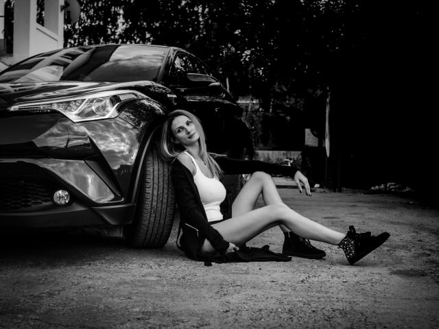Mila McMillan, ass visible, appuyée contre une voiture, portant un haut blanc, une veste noire et des bottes noires, les jambes croisées, regardant vers le haut, photo en noir et blanc..