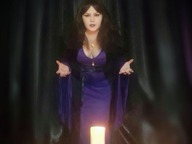 KaryQueen mit dunklem Haar, die ein violettes Kleid, eine schwarze Jacke und eine Halskette trägt, steht mit ausgestreckten Armen vor einer leuchtenden Kerze und wirkt ernst. Ihre Brüste sind sichtbar..