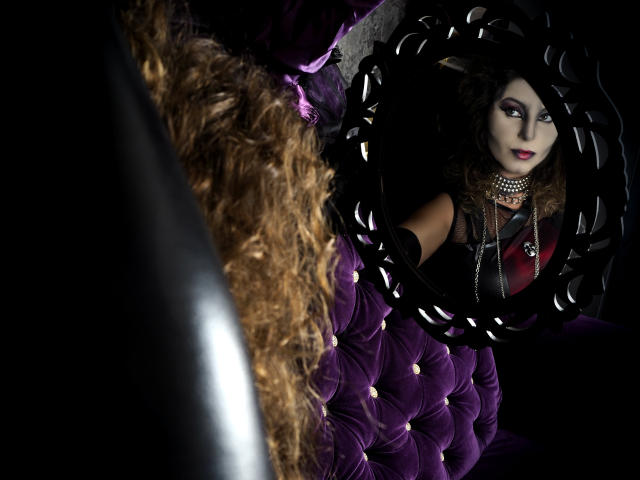 VelvetManttis with curly brown hair, black dress, and red necklace, reflected in a black, ornate mirror, sitting on a purple, tufted velvet couch. Boobs visible in reflection.