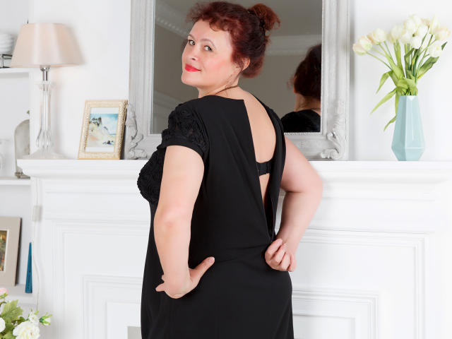 BerryChic, con cabello rojo rizado, sonriendo, vestida con un vestido negro, mostrando su espalda, con las manos en las nalgas, reflejada en un espejo, flores blancas en un jarrón azul..