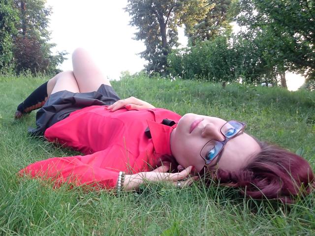 MoonBlossom liegt auf dem Gras, die Beine angezogen, und trägt ein rotes Hemd, einen schwarzen Rock und eine Brille. Sie hat rotes Haar und ein süßes Gesicht, wobei ihr Hintern leicht angehoben ist..