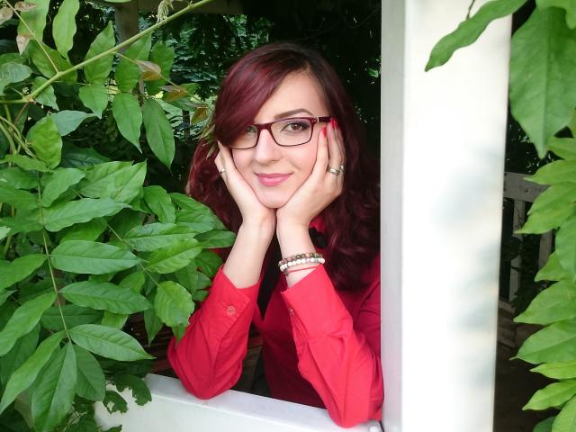 MoonBlossom with red hair, glasses, and red shirt, resting her chin on her hands, surrounded by green leaves, wearing a bracelet and ring, smiling subtly.