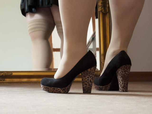 As pernas de BonnieAngel, com saltos pretos estampados com padrão de leopardo e plataformas estampadas com o mesmo padrão, estão em frente a um espelho que reflete suas coxas pálidas e a saia preta..