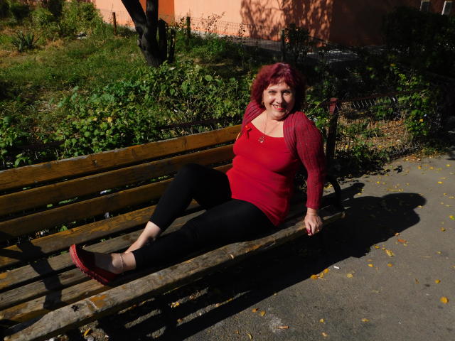 SexyEllaForYou is een mollige vrouw met rood haar, een rode top, zwarte broek en rode schoenen, die op een houten bank zit, lachend, met gespreide benen..
