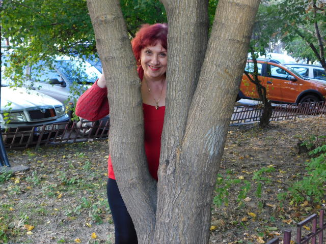 SexyEllaForYou with red curly hair and red sweater, standing behind thick tree trunks, smiling, holding her hair. Car and fence in background. Autumn leaves on ground.