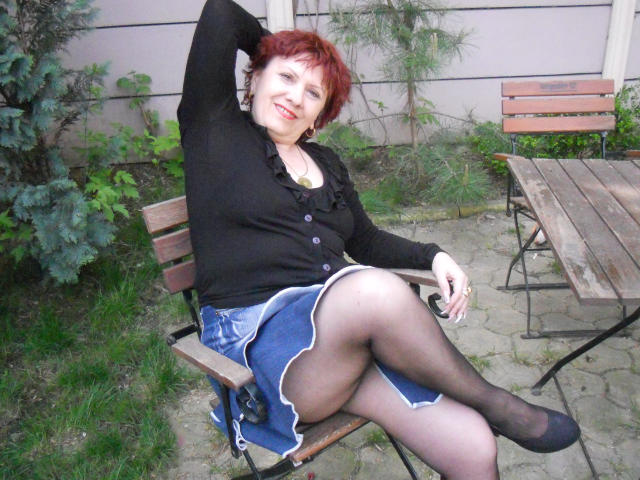 SexyEllaForYou con cabello rojo, cárdigan negro, falda azul, medias negras y zapatos negros, sentada en un banco de madera, con el brazo levantado, sonriendo, con las piernas cruzadas, mostrando su trasero..