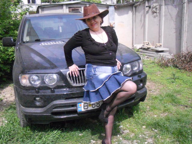 SexyEllaForYou, con los pechos a la vista, usando un sombrero marrón, una blusa negra, una falda azul y tacones negros, se apoya en un coche negro, mostrando su vulva a través de su falda..