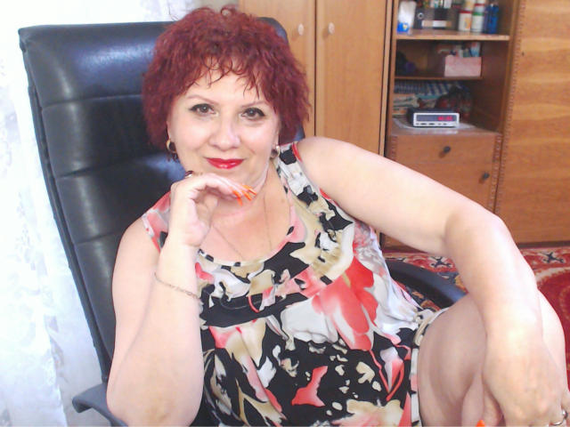 SexyEllaForYou with curly red hair, red lipstick, and a colorful floral dress, sitting in a black chair, touching her chin with orange nails, smiling gently.