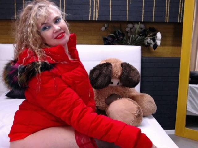 LadyMariahX with curly blonde hair, red puffy jacket, and red lipstick, sitting on a bed with a brown teddy bear, smiling at the camera, showing her white thighs.