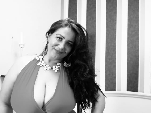 Black-and-white photo of LexyRose with long dark hair, smiling, wearing a halter top showing big tits, and a floral necklace. Her boobs are prominent, and she's looking cute.