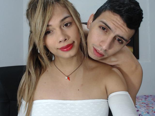 ValeriTSCpl, com maria-chiquinhas loiras, batom vermelho e uma blusa branca com um ombro descoberto, tem os braços em volta de ValeriTSCpl, que está sem camisa, tem cabelo escuro e uma expressão divertida..