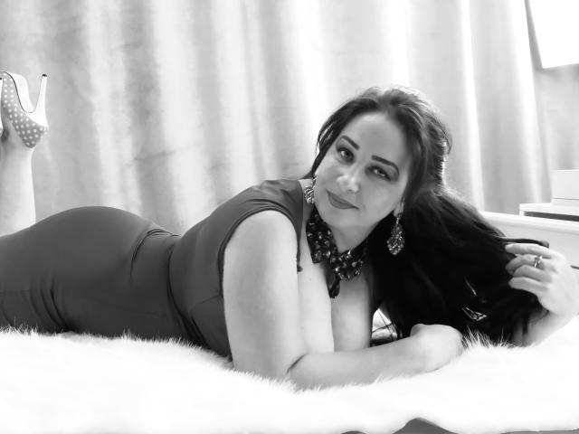 LexyRose lies on a fluffy rug, wearing a tight dress that highlights her ass, with long dark hair, earrings, and a necklace, in a black-and-white photo.